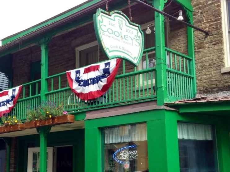 The hanging shield sign at Cooley's reading Est. 2004, Cooley's Stone House Tavern, Food and Spirits, above the green porch and the open front door