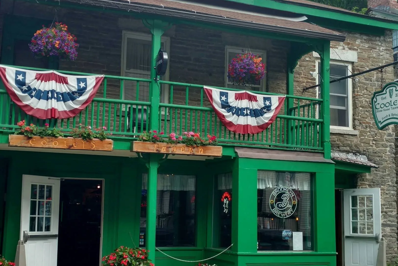 The green porch and fieldstone front of Cooley's Stone House Tavern on Pioneer Street, Cooperstown, with bunting on the balcony rail and carved Cooleys Tavern flower boxes